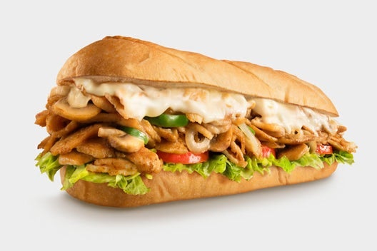 Chicken Philly Cheesesteak from Charleys Cheesesteaks against a white background. This is make with grilled chicken, peppers, and onions with provolone on a toasted roll. This is served with lettuce, tomato, mayo, and pickle. Philly chicken cheesesteak near me.