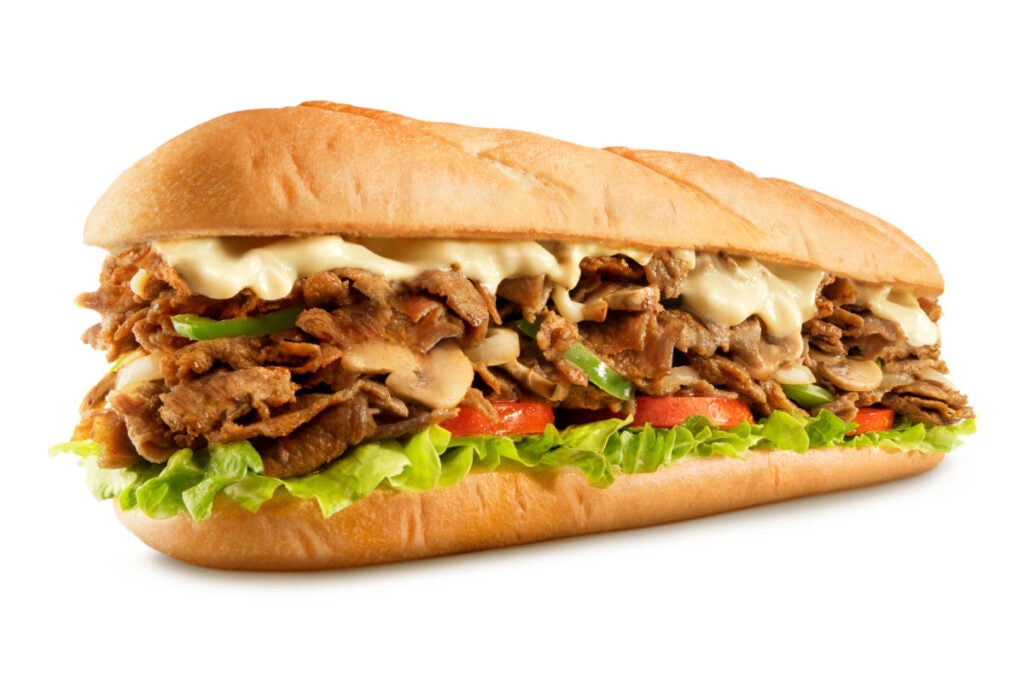 Steak Philly Cheesesteak from Charleys Cheesesteaks against a white background. The Steak Philly Cheesesteak is made with melted provolone cheese, grilled steak, onions, and peppers on a toasted roll, then topped with lettuce, tomato, mayo, and pickle.