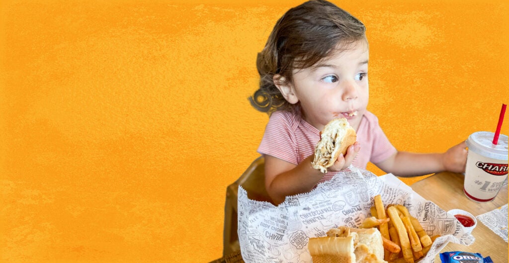 Child eating a Kids Meal from Charleys Cheesesteaks, including a cheesesteak, fries, and Oreo cookie, against an orange background.