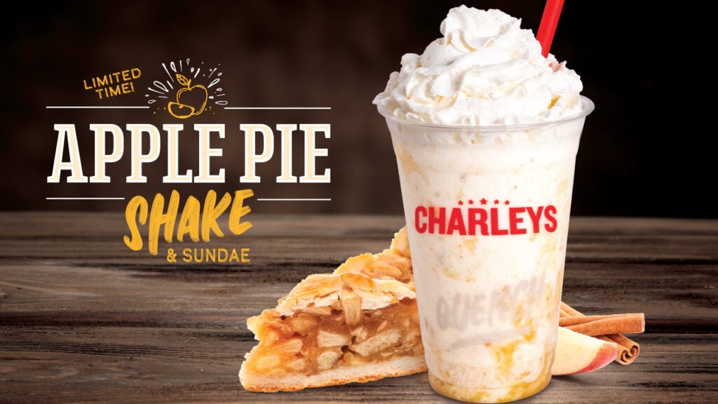 The Apple Pie Shake with whipped cream, sitting on a table with a slice of apple pie with a cinnamon stick.