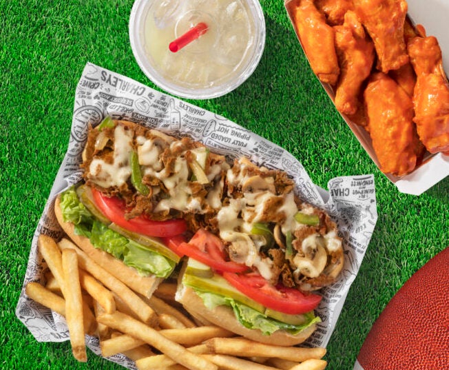 A cheesesteak with french fries, wings, and a lemonade from Charleys Cheesesteaks sitting on turf with a football nearby.