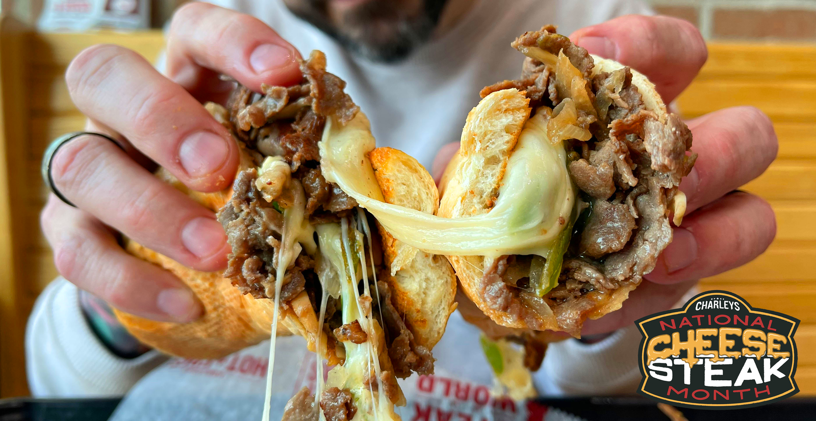 A person holding a cheesy cheesesteak from Charleys with a logo in the corner that says National Cheesesteak Month.