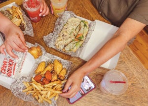 People reaching across a table with cheesesteaks, wings, fries, lemonades, and the Charleys Rewards app at a Charleys Cheesesteaks and Wings.