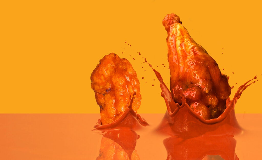 Two chicken wings dunking in a sea of buffalo sauce. Celebrate National Chicken Wing Day with Charleys Cheesesteaks and Wings.